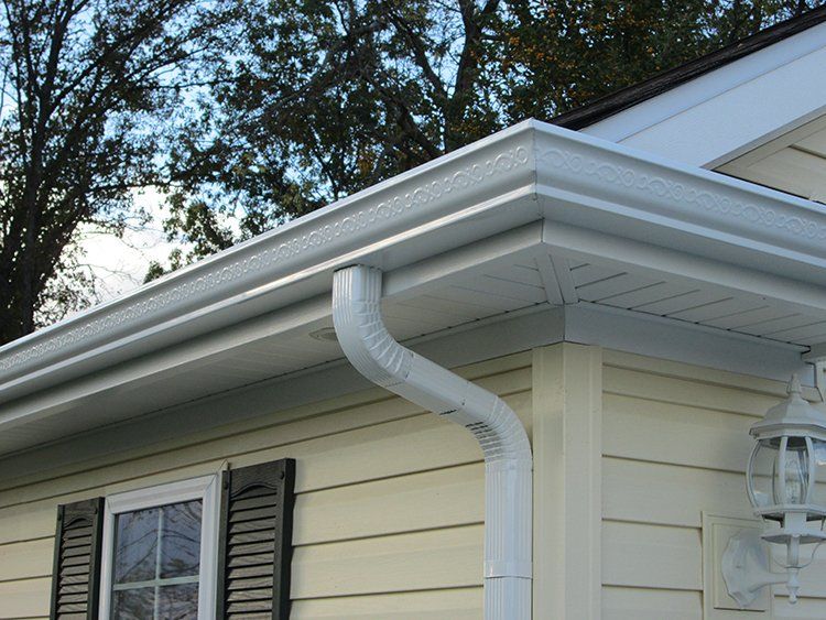 White house roof eave with a curved gutter and downspout above a window with dark shutters
