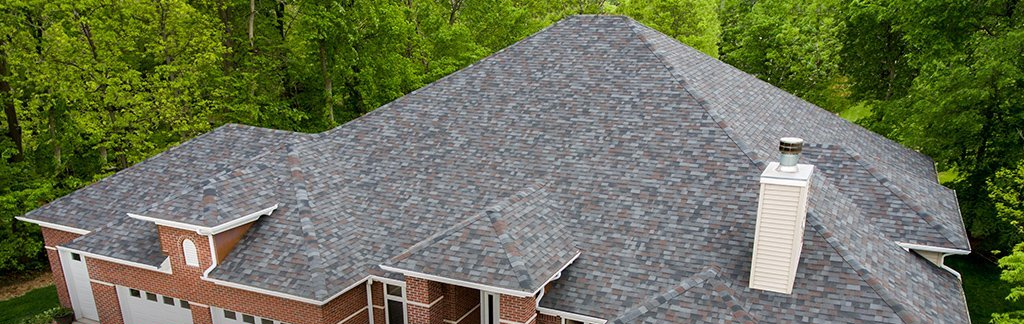 Aerial view of a brick house with a gray shingled roof surrounded by green trees