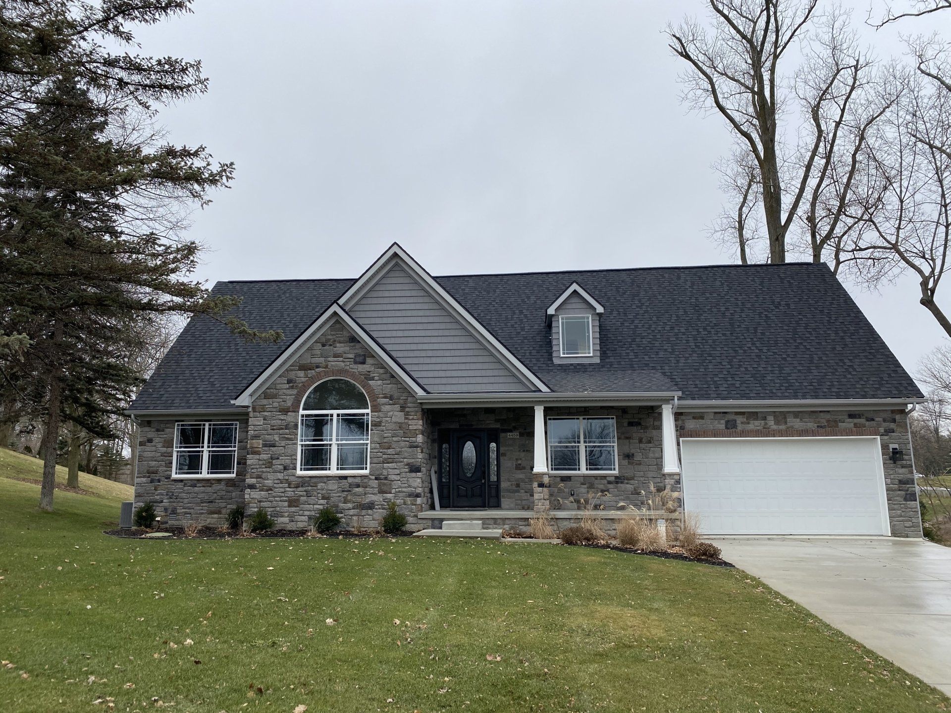 Stone house with a dark roof, front lawn, and white garage under an overcast sky.