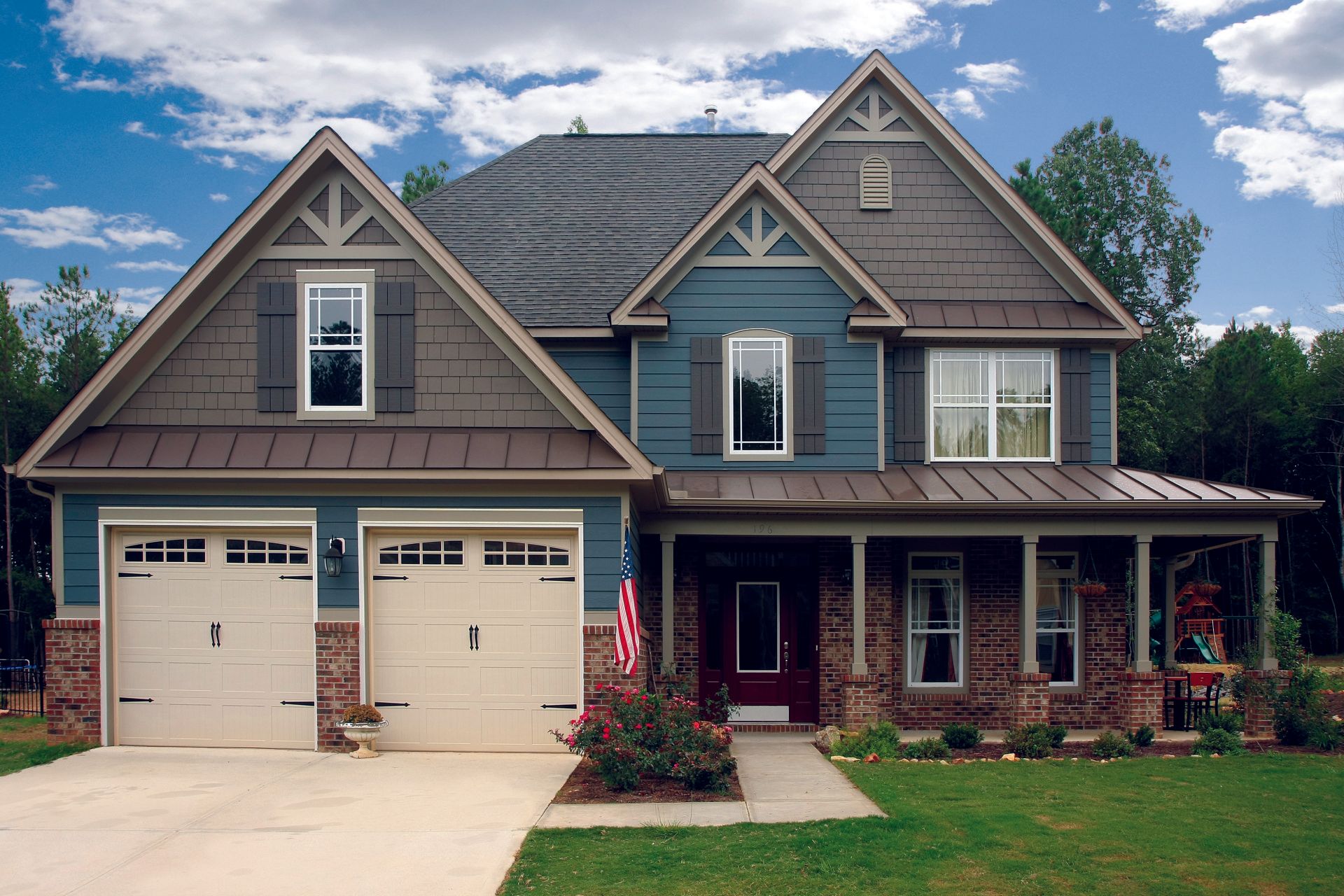 Suburban two-story house with gray siding, stone porch, two-car garage, and a U.S. flag by the entry