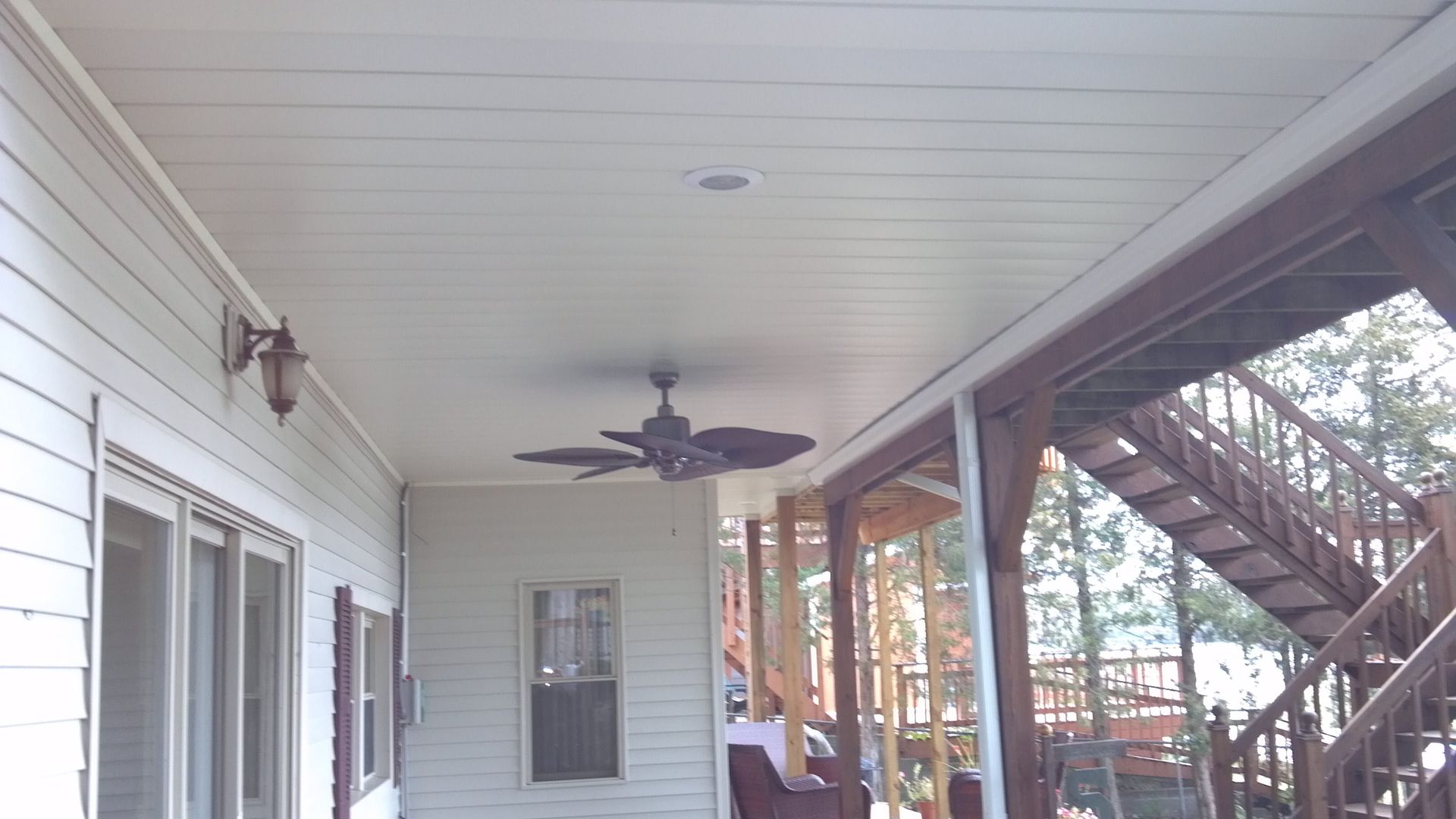 Covered porch with white siding, ceiling fan, windows, and screened stairs overlooking a wooded yard