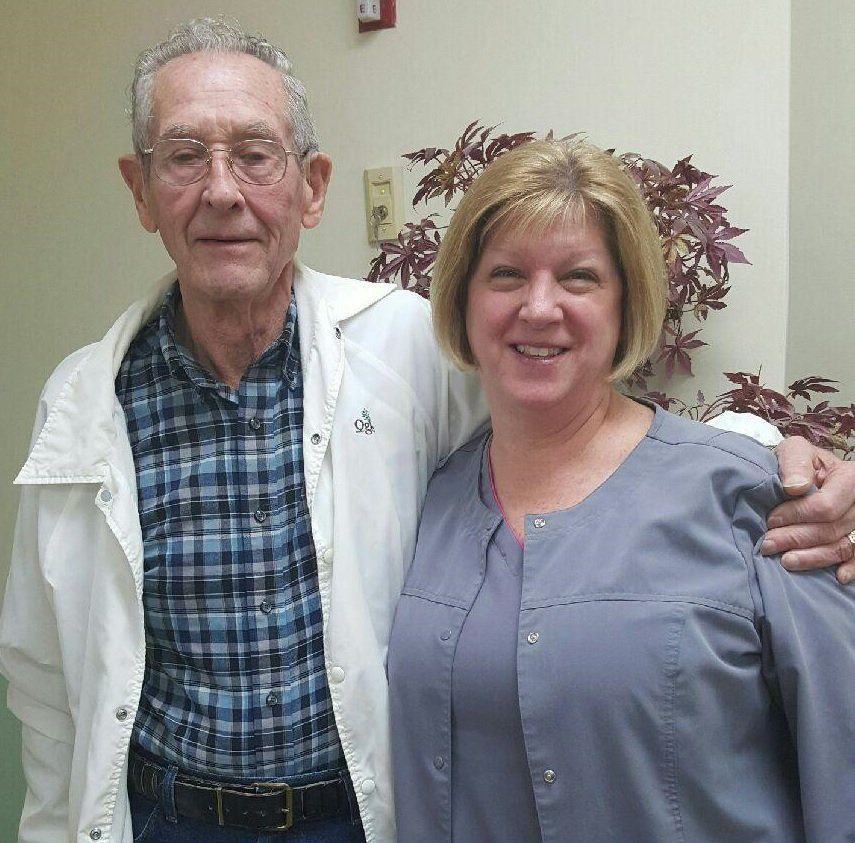 Two people smiling indoors, standing close together in front of a plant.