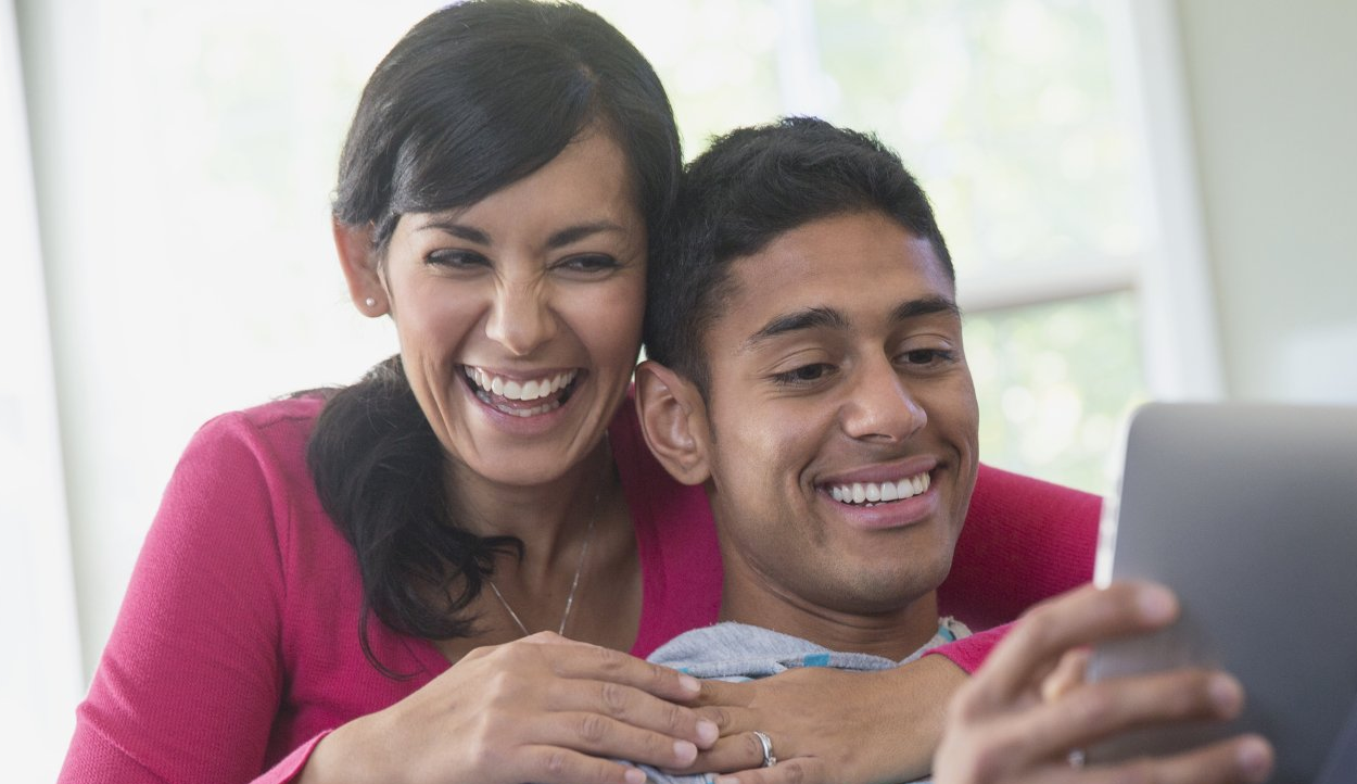 Smiling couple taking a selfie on a laptop, with one person hugging the other from behind.