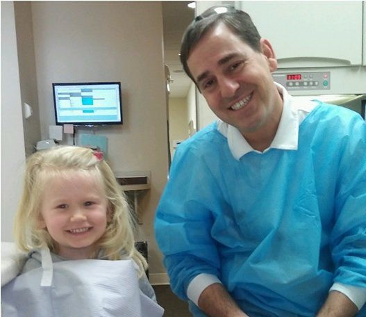 Smiling man in blue medical gown stands beside a smiling child in a dental office
