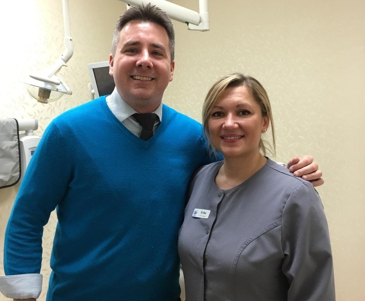 Two smiling coworkers standing together in a hallway, one in a blue sweater and one in gray scrubs.