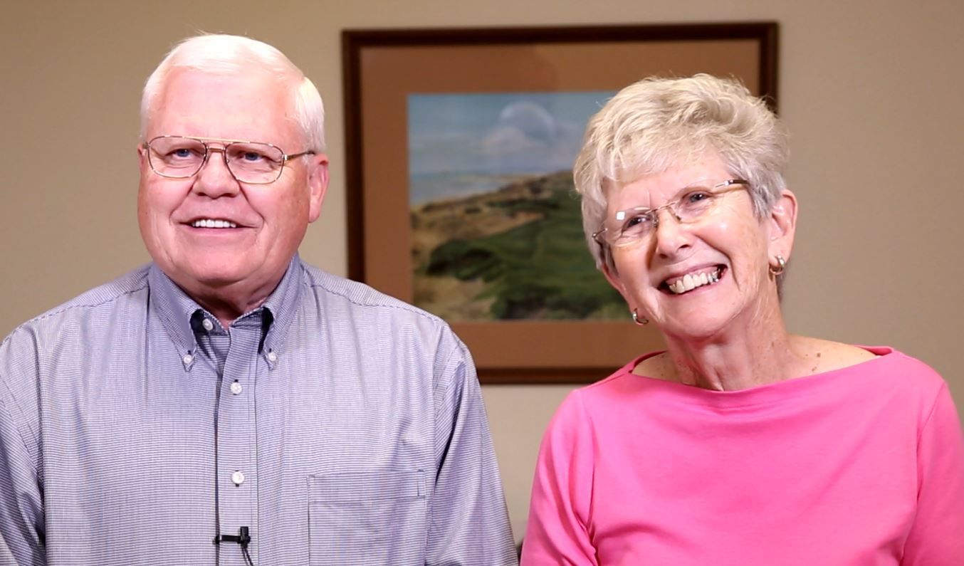 Two smiling people seated indoors, one in a blue shirt and one in a pink top, with a framed landscape behind them