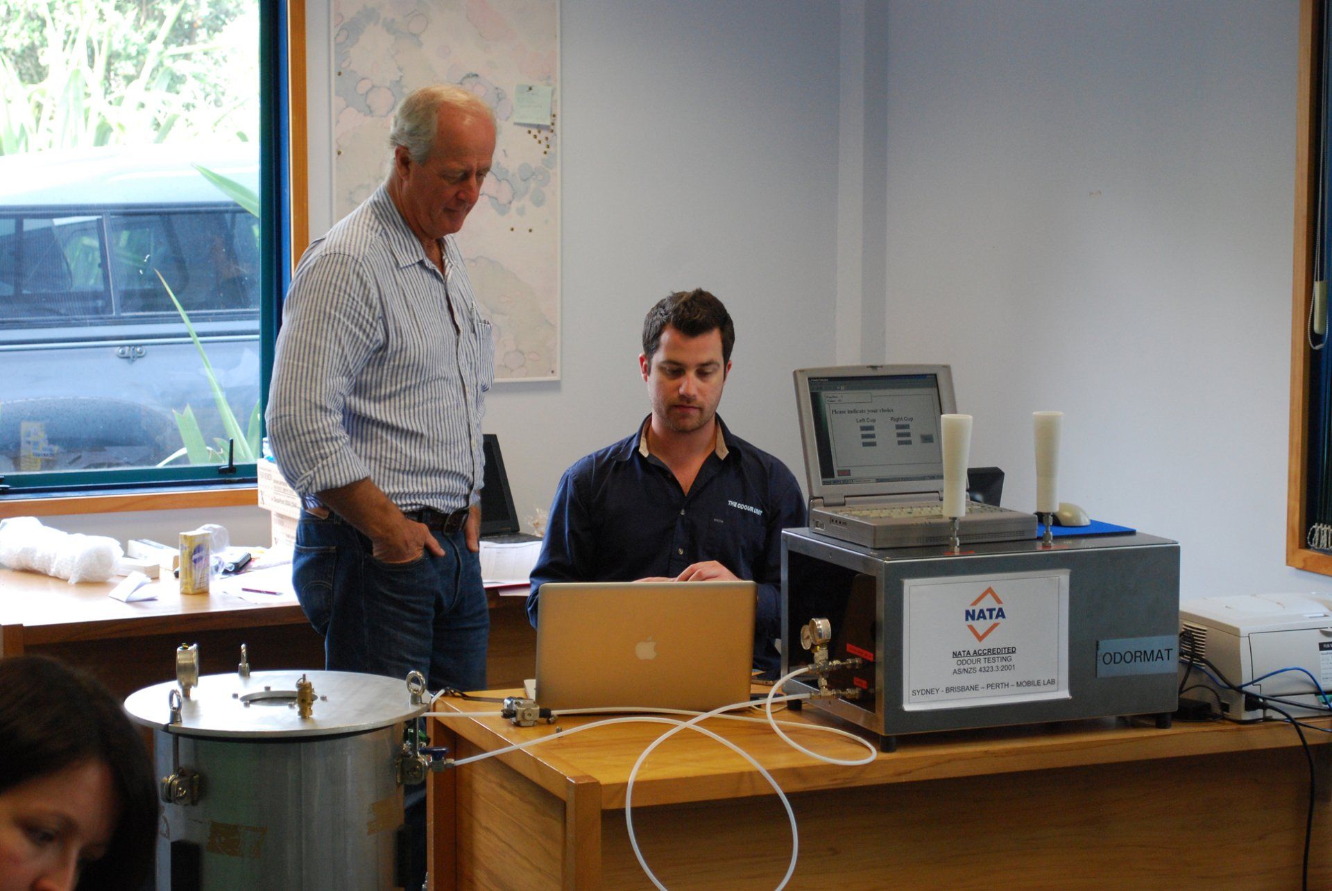 Two men working with a machine and laptop at a table in a lab-like setting. One is standing, looking on.