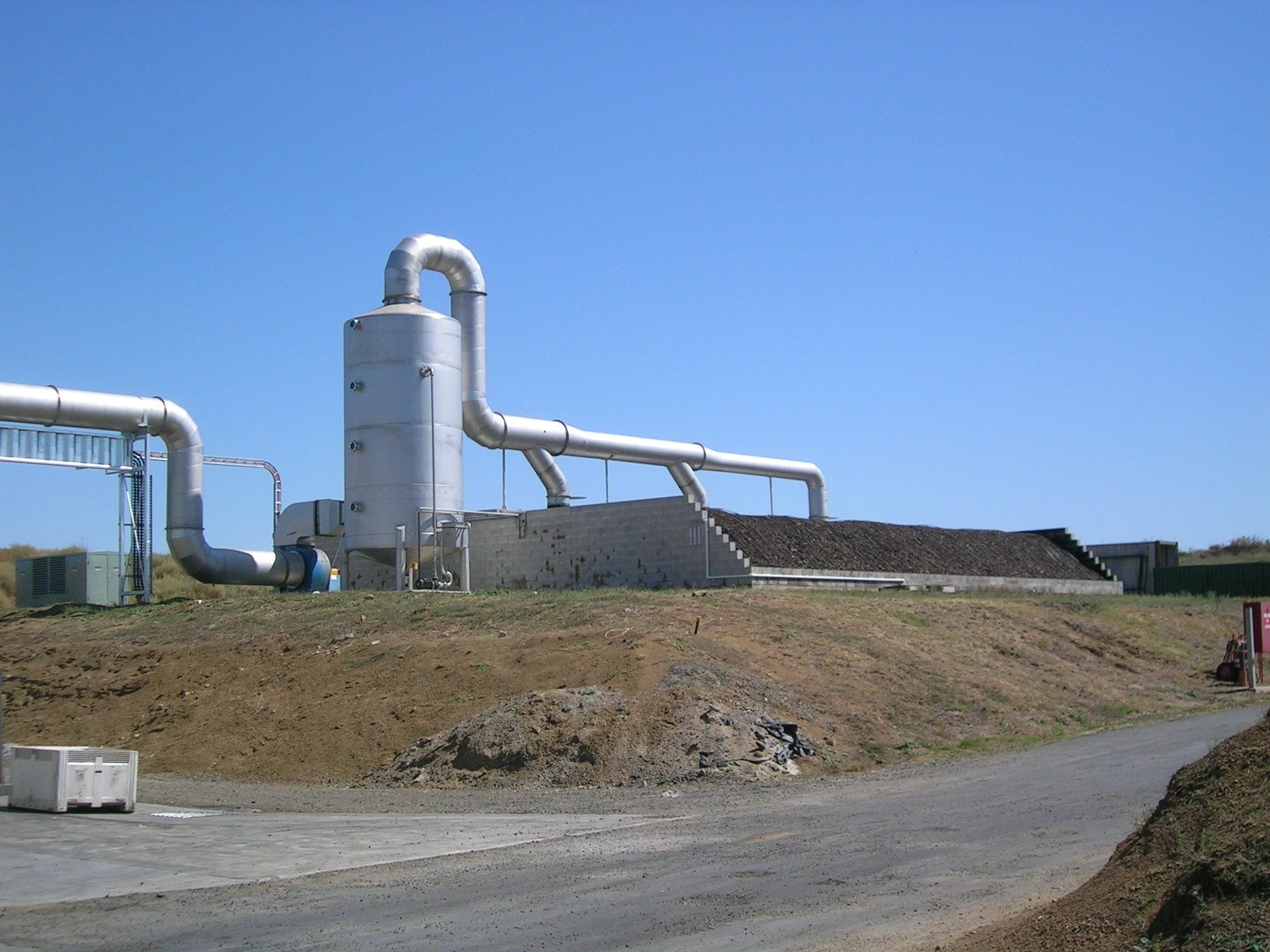 Industrial facility with pipes, tanks, and a pile of organic material against a blue sky.