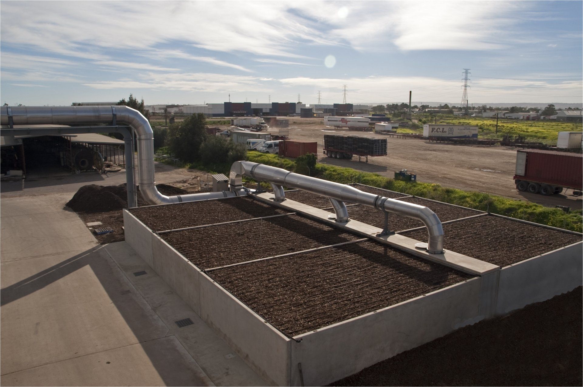 Rooftop with metal pipes and a raised bed covered in brown material, with a distant industrial landscape.