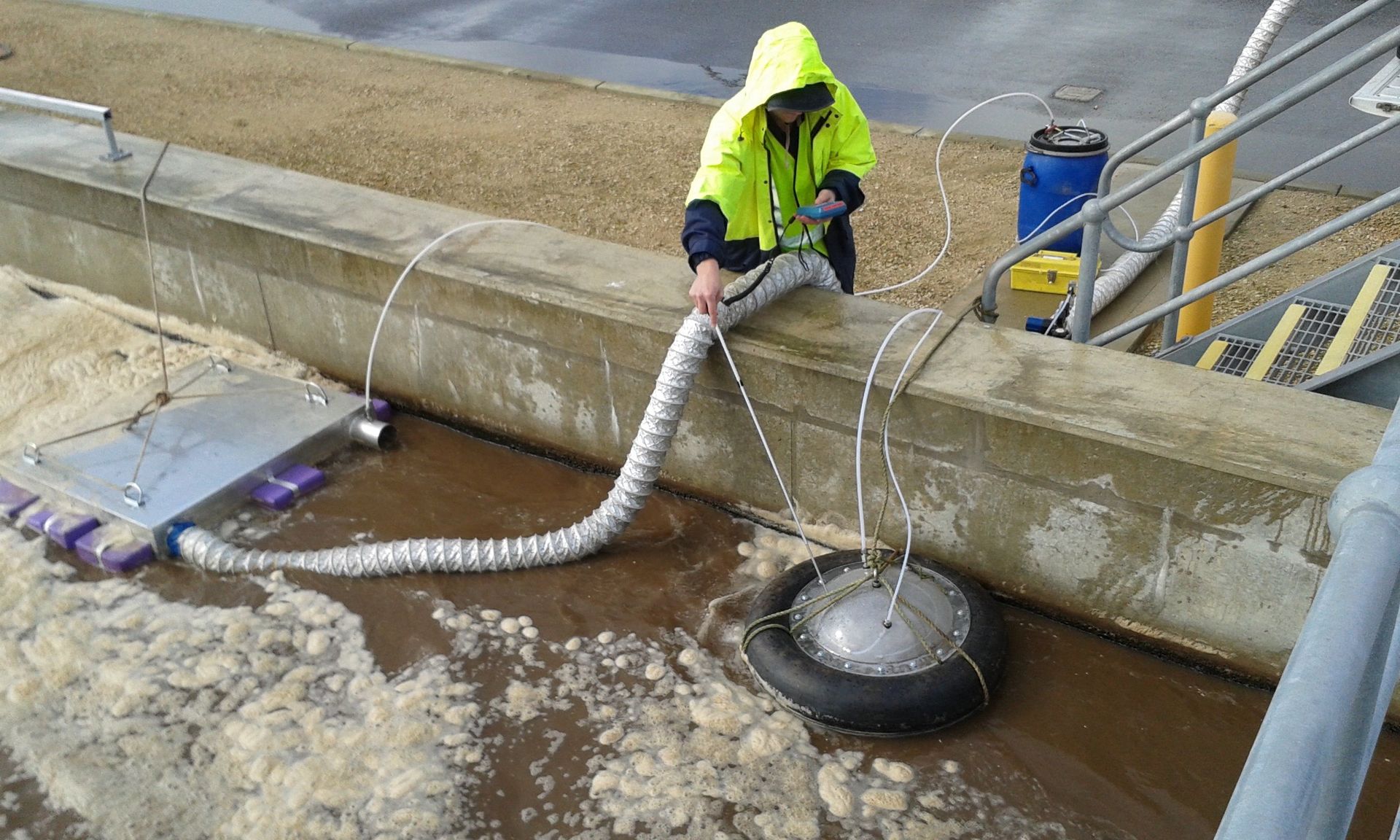 Person in safety gear using a hose connected to a device in brown, foamy water at an industrial site.