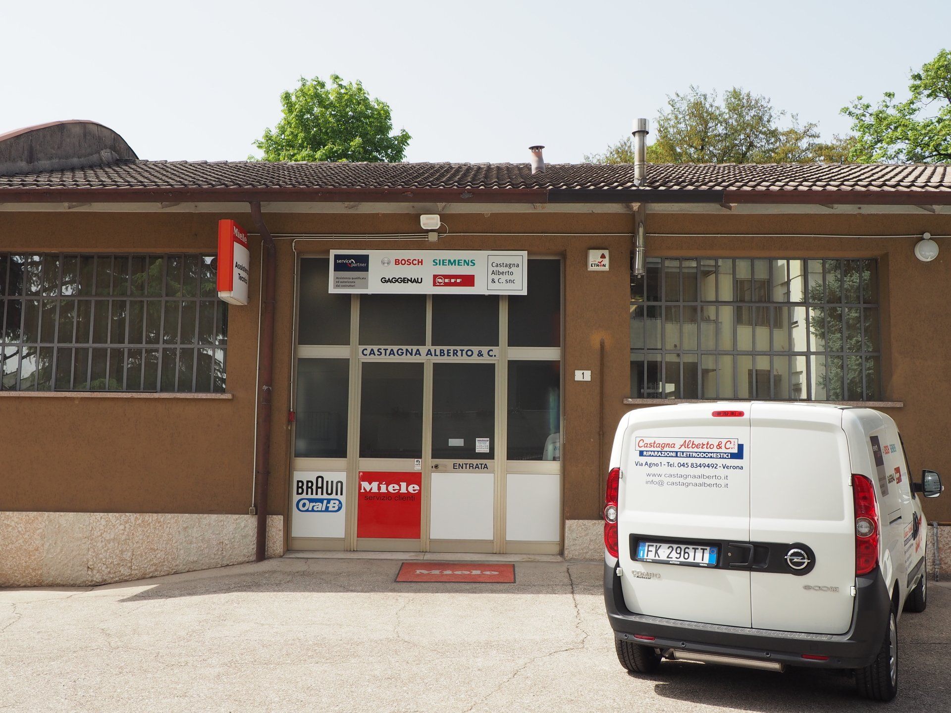 Esterno di un piccolo negozio con ingresso in vetro, finestre con sbarre e un furgone bianco parcheggiato davanti.