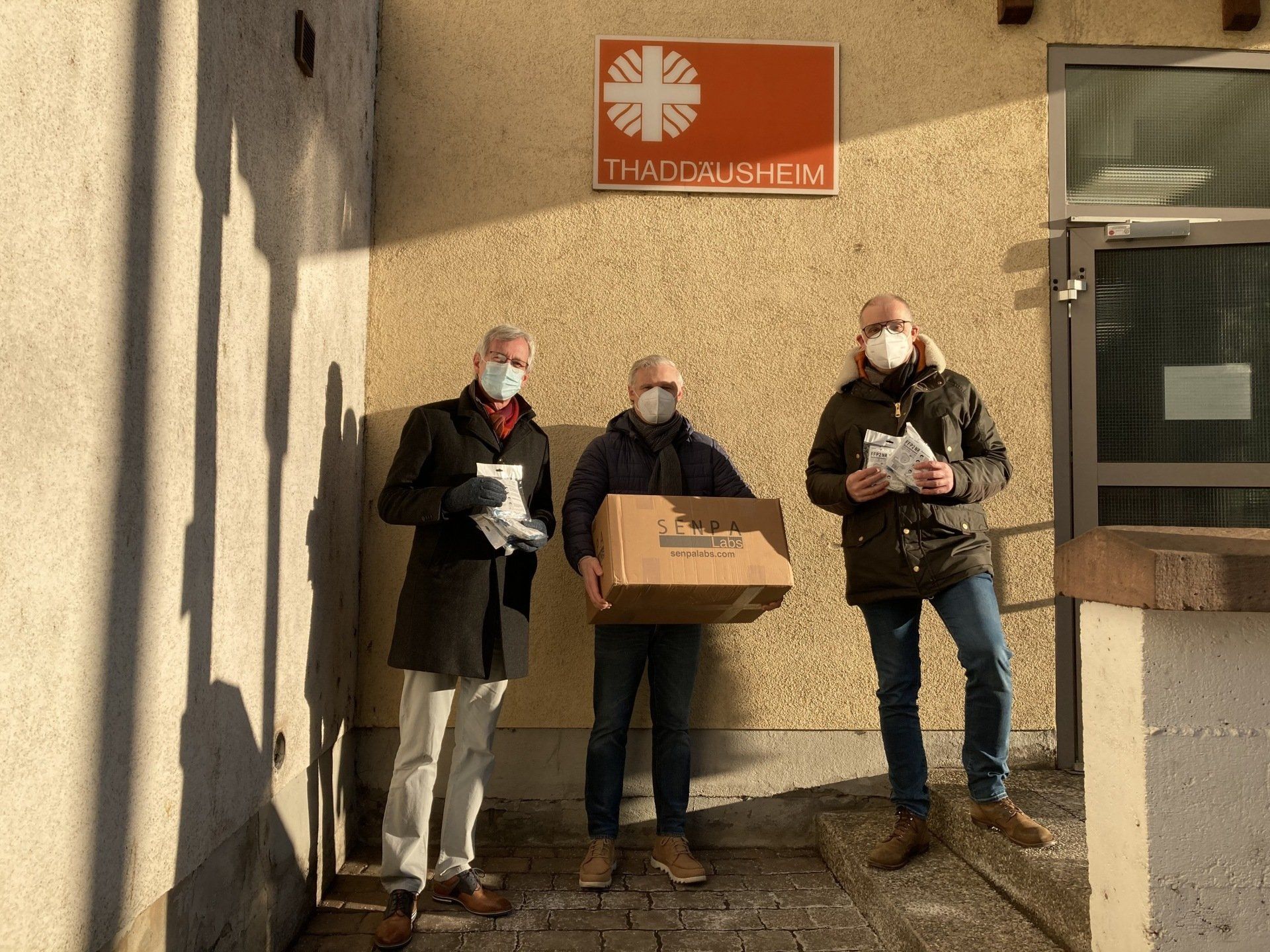 Drei Männer mit Gesichtsmasken stehen vor einem Gebäude mit einem Schild mit der Aufschrift „Caritas“