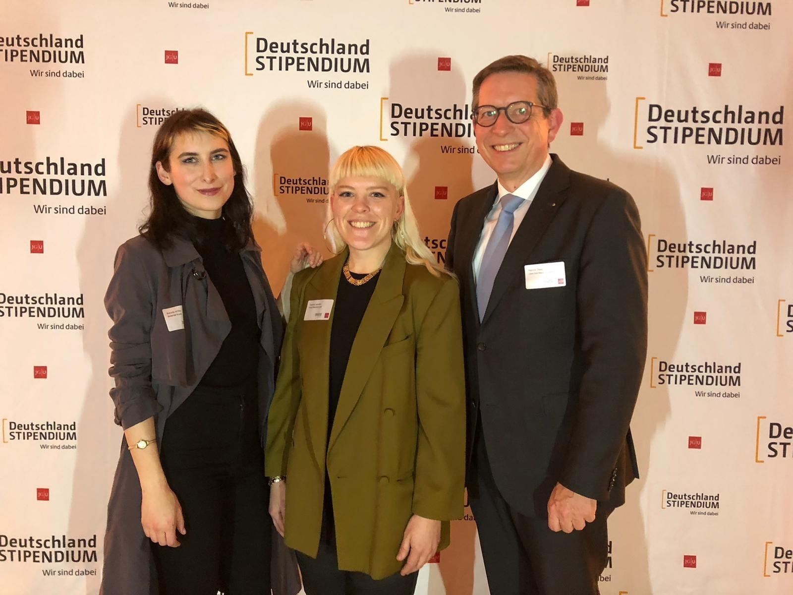 Drei Personen posieren für ein Foto vor einer Wand mit der Aufschrift „Deutschland Stipendium“