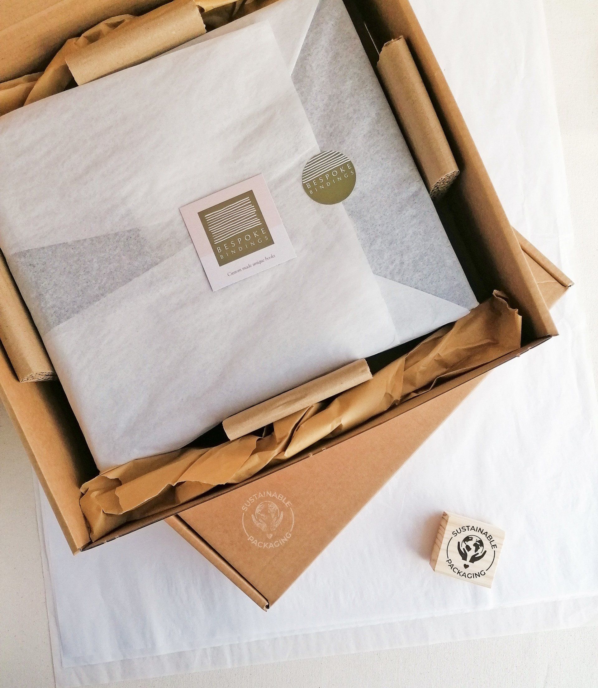 The image shows a beautifully wrapped handmade book in recycled white tissue paper within brown sustainable packaging boxes and a Bespoke Bindings business card on top with the green logo. The open box is stacked on top of another box with a stamp saying Sustainable Packaging.
