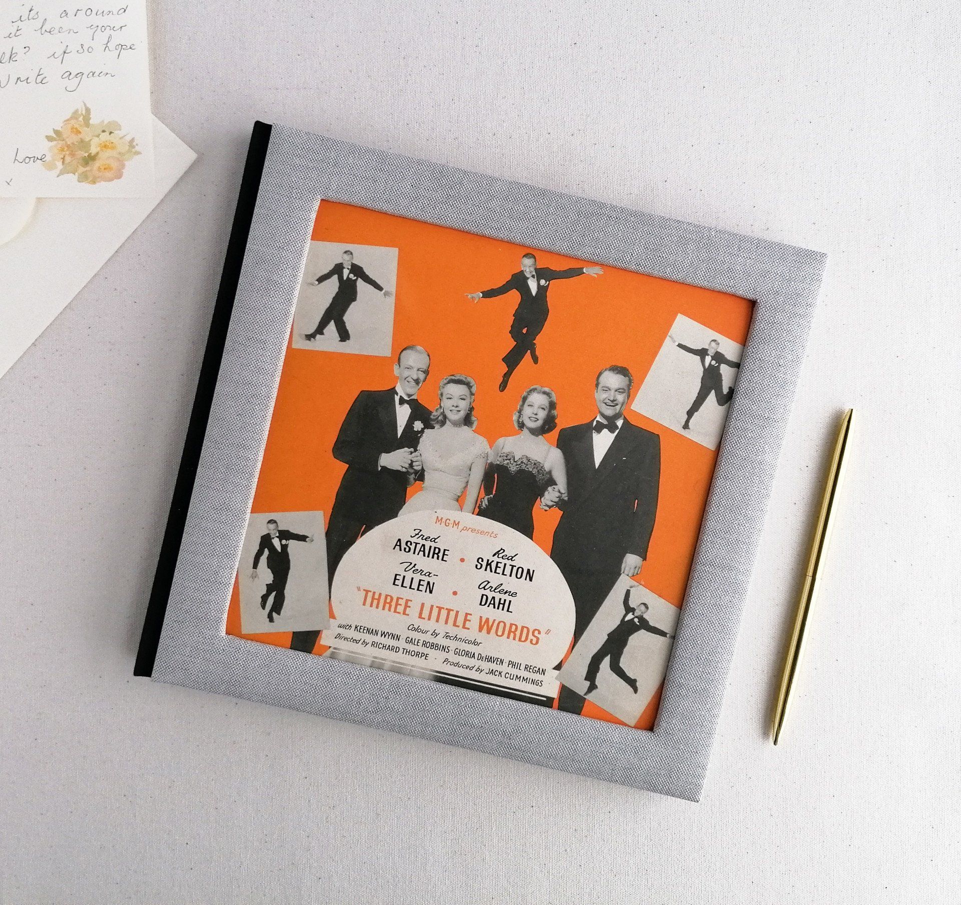 An image of a large grey linen scrap book with padded covers and a cut out on the front cover with a vintage sheet music image. The image is orange in the background with black white and grey photos of 2 ladies and 2 men at the forefront. The sheet music is advertising 3 little words with Frank Sinatra and Frank Sinatra is dancing in 5 places within the background. The scrapbook is 10 and a half inches square in size and is made as a hardback book. The spine is black which contrasts against the grey cover and orange vintage sheet music image. The album is laid on a white background next to a handwritten letter on floral notepaper.