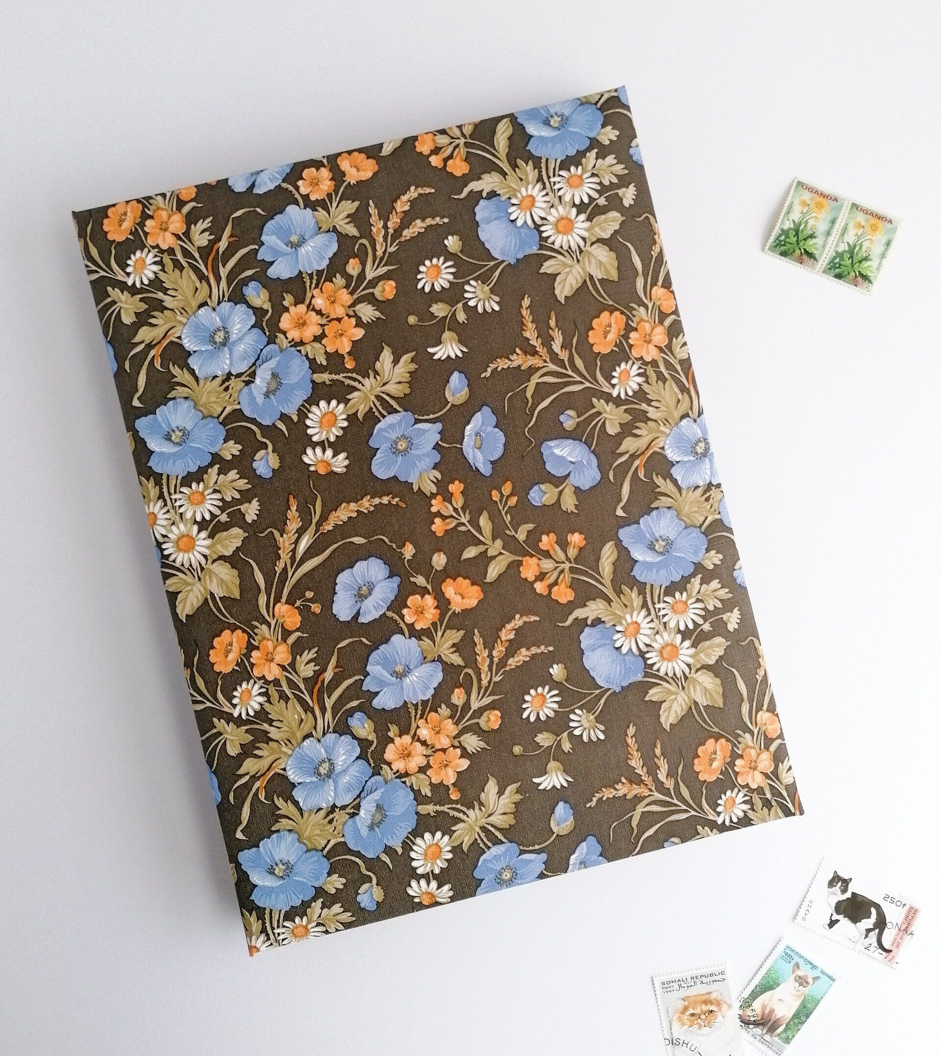 The image shows a portrait A4 sized custom made stamp book album. The covers are padded with a decorative vintage floral wallpaper. The flowers are light blue olive green and orange on a soft black background. There are stamps with images of cats on the table next to the album.