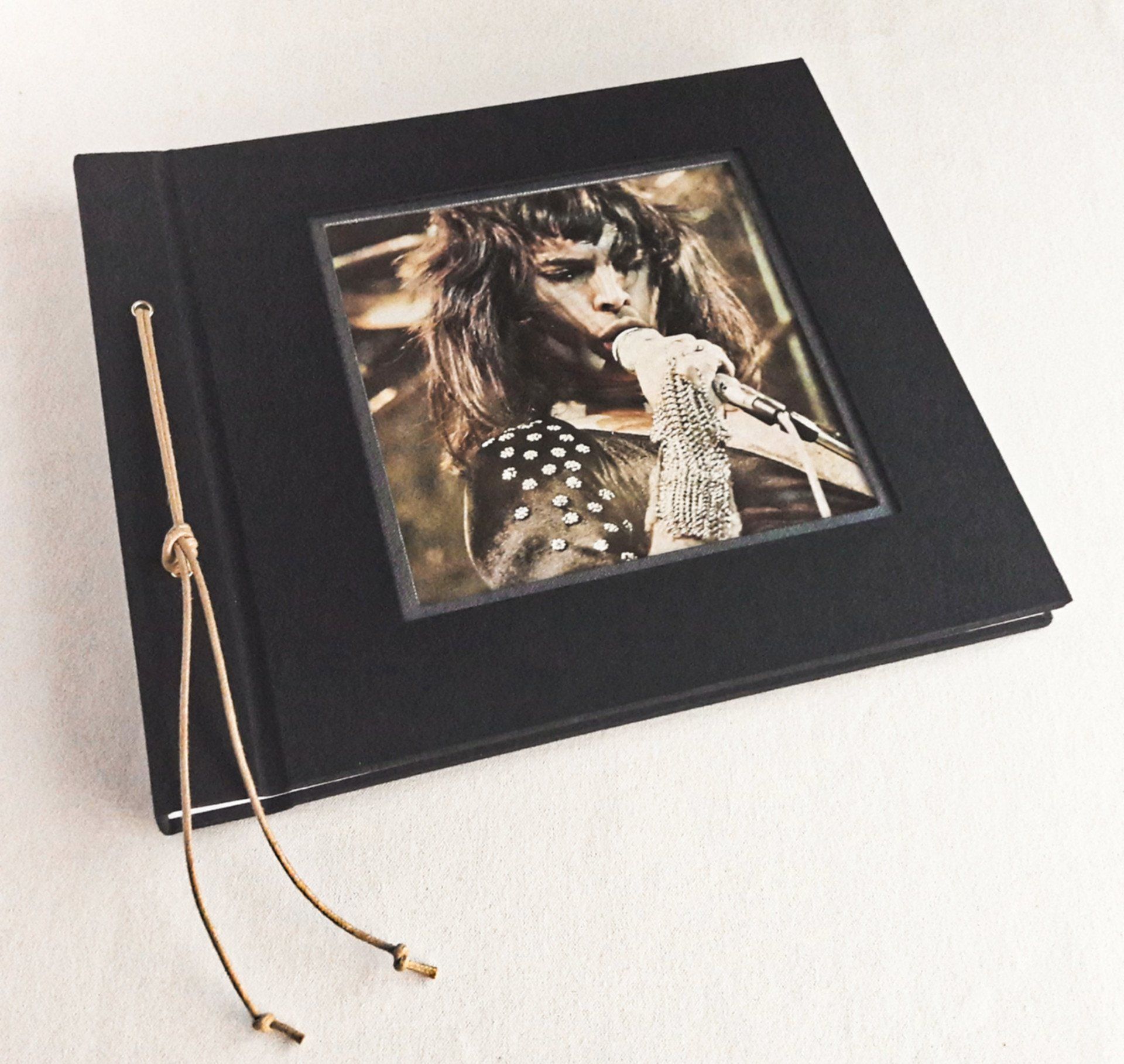 The image shows a bespoke landscape photo album covered in black book cloth with a vintage Freddie Mercury image from a magazine designed within a cut out on the front cover.
