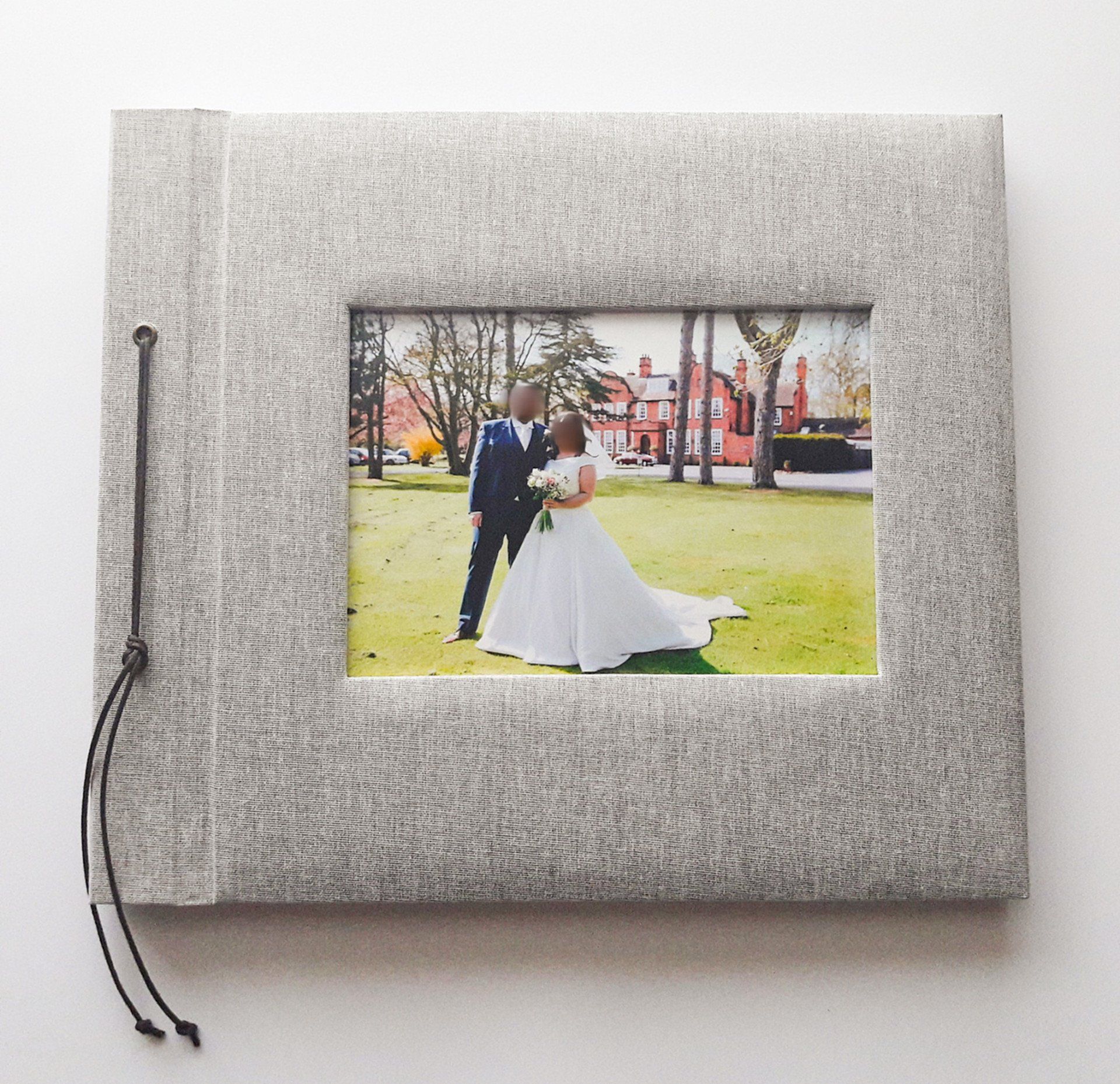 The image shows a light Grey linen large album with padded covers. The album is bound with black cord and knotted to the front. There is a photo within a cut out section of the front cover which shows a couple getting married.