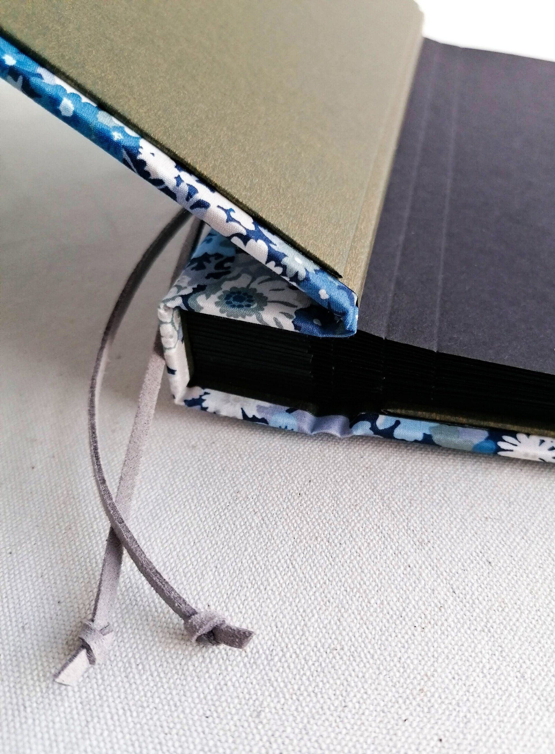 The image shows the detail of a large landscape photo album which is opened at the first page. The inside of the album cover is gold. The album has black pages and these are scored to enable the pages to open with ease. The album has a grey suede cord threaded through to bind the cover and pages together. The decorative material on the cover is floral in light blue grey dark blue white and beige. The fabric is by Liberty of London. The album is laid on a white background.