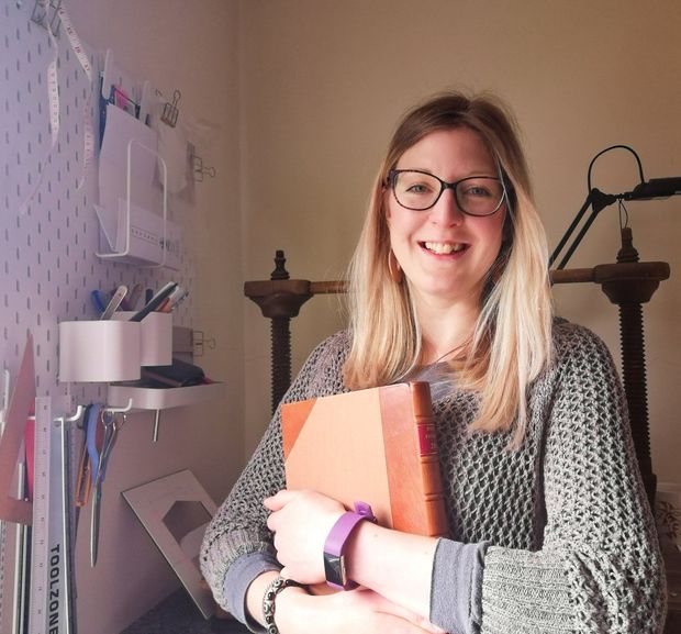 There is a woman called Holly who is the BookBinder of Bespoke Bindings. She is holding a book with a brown leather spine and cloth covered sides that she has handmade. She stands in front of a large Victorian wooden sewing frame in her studio with her tools hanging on the wall beside her. Holly is smiling and she has long blonde hair and wears glasses. She wears a brown open weave jumper.