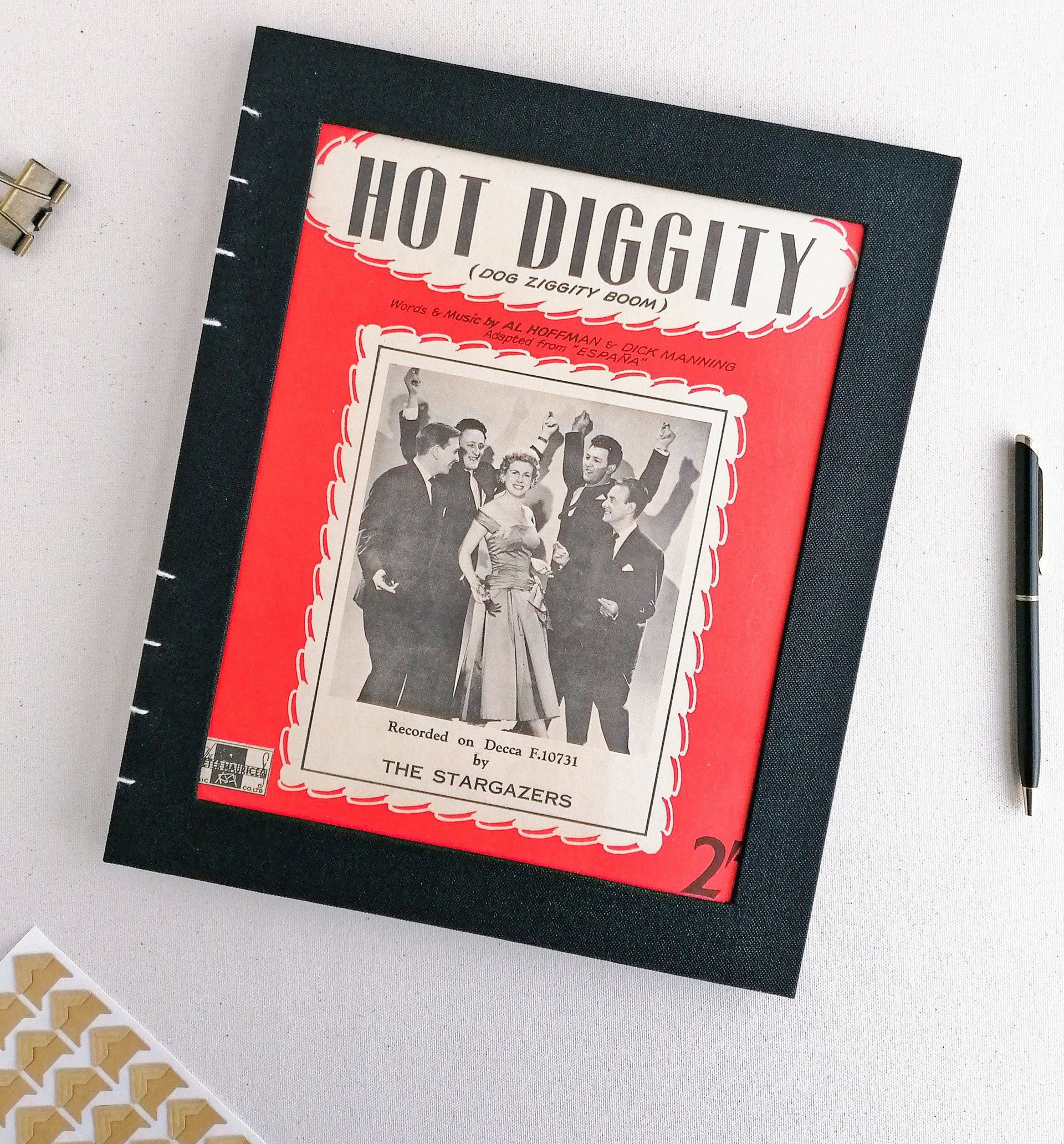 An image of a large black linen portrait scrap book with a cut out on the front cover. The image within the cut out is taken from vintage sheet music which says Hot Diggity. The black and white image shows 4 men dressed in black suits dancing around a lady in a satin cocktail party dress. The photo is from between the 1940's and 1950's. The photo has a bright red border which contrasts nicely against the black linen cover. The spine is sewn and the pages are exposed on the spine. This is called a Coptic style of traditional bookbinding. The album is laid on a white background next to some small brown craft card photo corners to be used in a scrapbook.