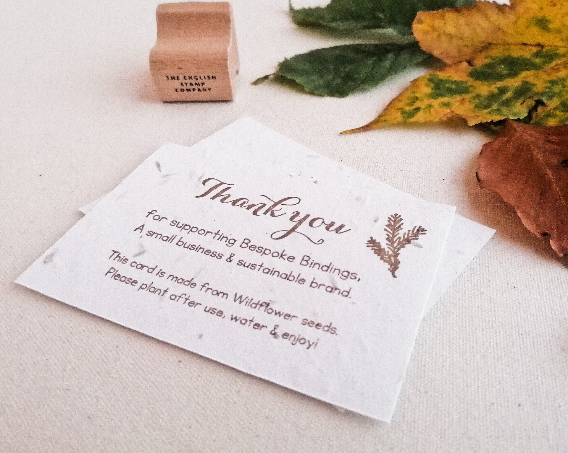 The image shows a thank you card from Bespoke bindings which is given out with every order. The card is cream and made from handmade paper pulp with seeds. The cards are stamped with the message Thank you for supporting bespoke bindings a small business and sustainable brand this card is made from wildflower seeds please plant after use and enjoy. The card is laid on a cream canvas table cloth with rustic autumnal leaves in green yellow and brown.