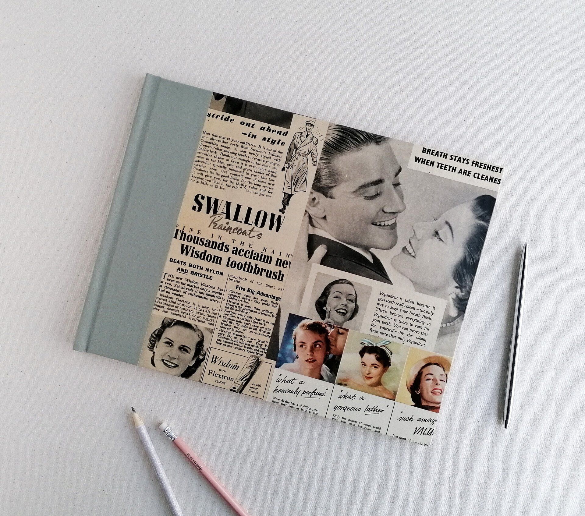 The image shows a large hardback slim sketchbook with a light green cloth covering the spine. The sides of the book on the front and back covers are decorated with vintage 1940's and 1950's advertisements for toothpaste clothing and beauty products. The advertisements show mens and womens faces smiling and the images are either in black and white or colour. The album is laid on a white background next to a light pink pencil and a light grey pencil.
