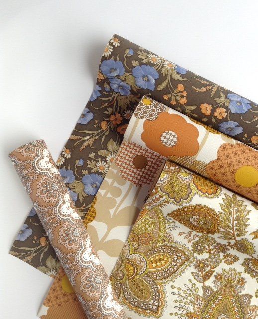An image of vintage retro patterned wallpaper in rolls. The top one is blue flowers olive green leaves and orange flowers on a soft black background. The second roll is modern orange mustard and chequered patterned brown flowers on a white background with light brown stalks. The third roll is a detailed paisley floral pattern of flowers and leaves in yellow light olive green and orange on a white background. They are all from the 1970's and 1960's era.