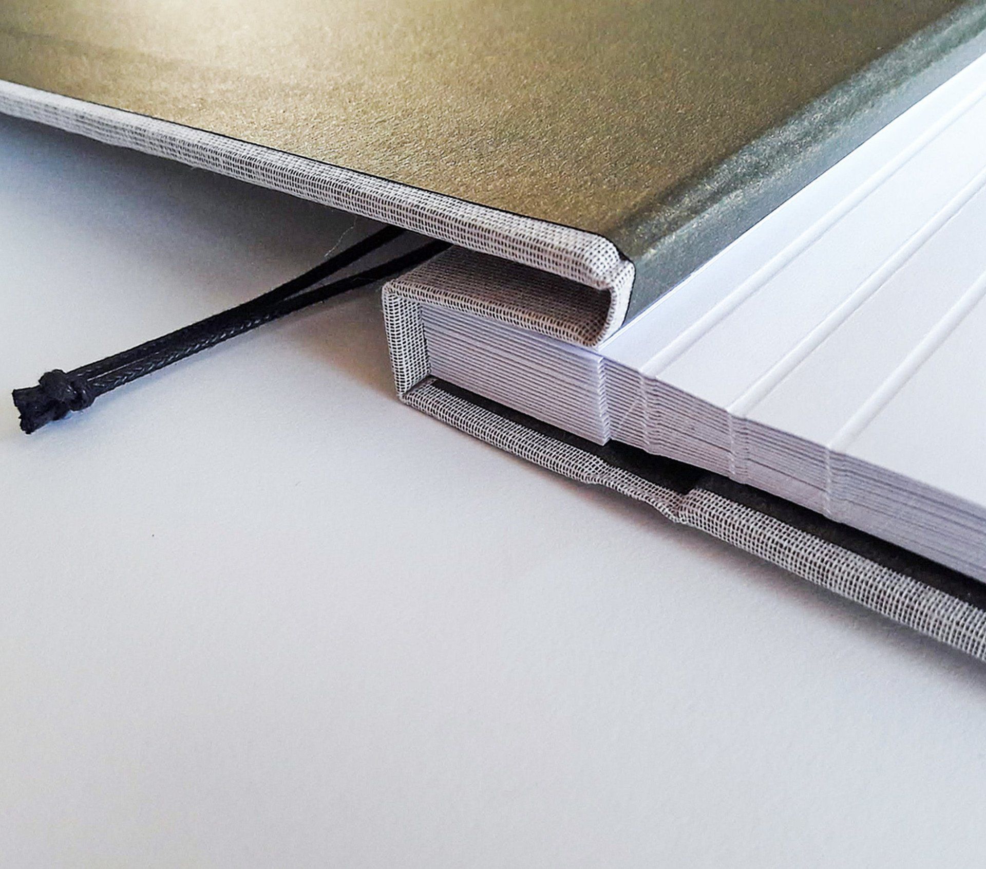 An image of a grey cotton landscape photo album. The image shows details of the album spine and neatly bound white page edges. The album is laid on a white background and the front cover is open. There are gold endpapers and white pages scored for ease of turning. The album has a black suede cord threaded through to bind the cover and pages together.