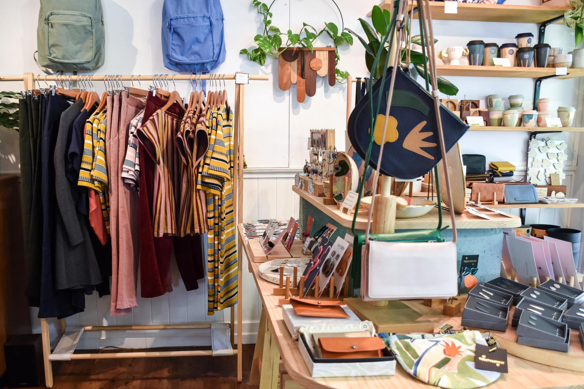 An image of a local boutique gift shop selling handmade goods by local artists and artisans. There is a rail of colourful clothing and 2 rucksacks hanging from the wall one blue and one green. There are some handmade leather hand bags and purses on display on a table colourful ceramic pots on shelves on the wall in the background.