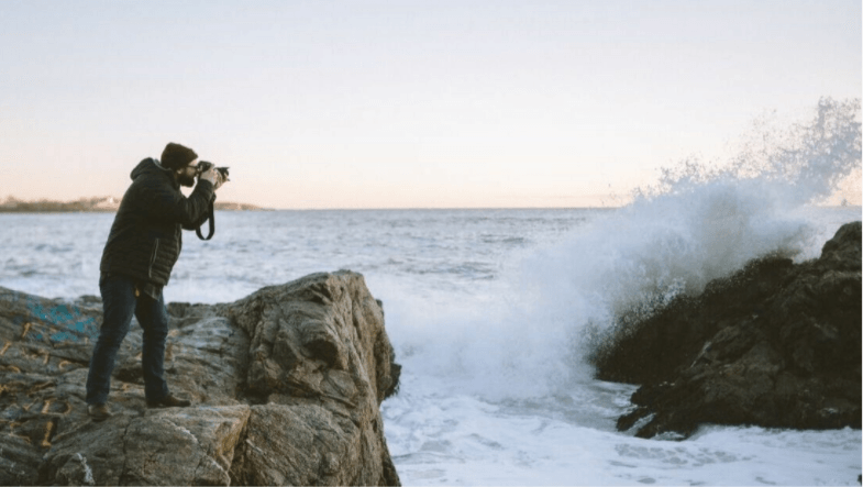 A man is standing on a rock taking a picture of the ocean.