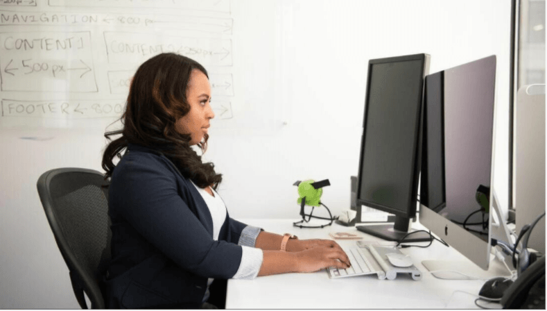 A woman is sitting at a desk in front of two computer monitors.