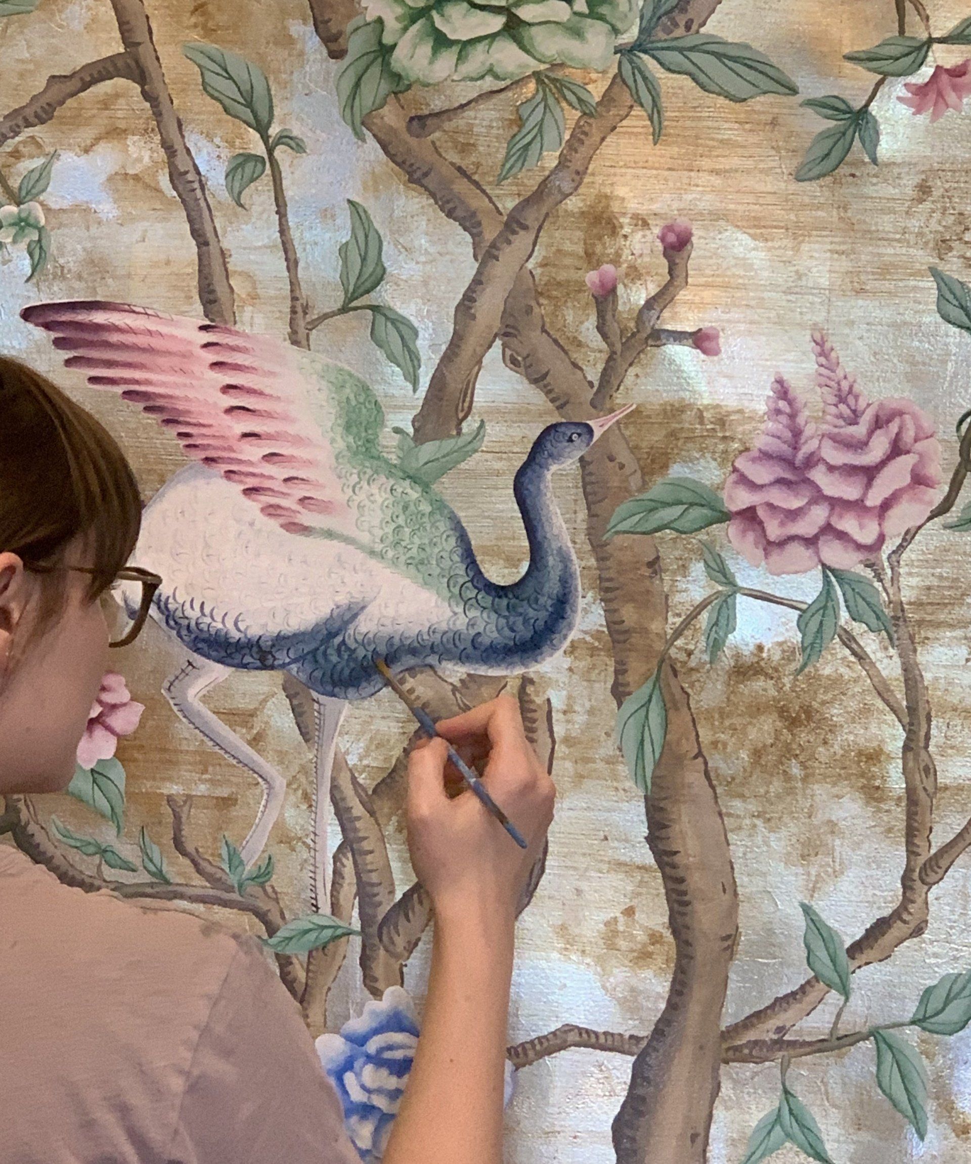 A woman is painting a bird on a wall with flowers.