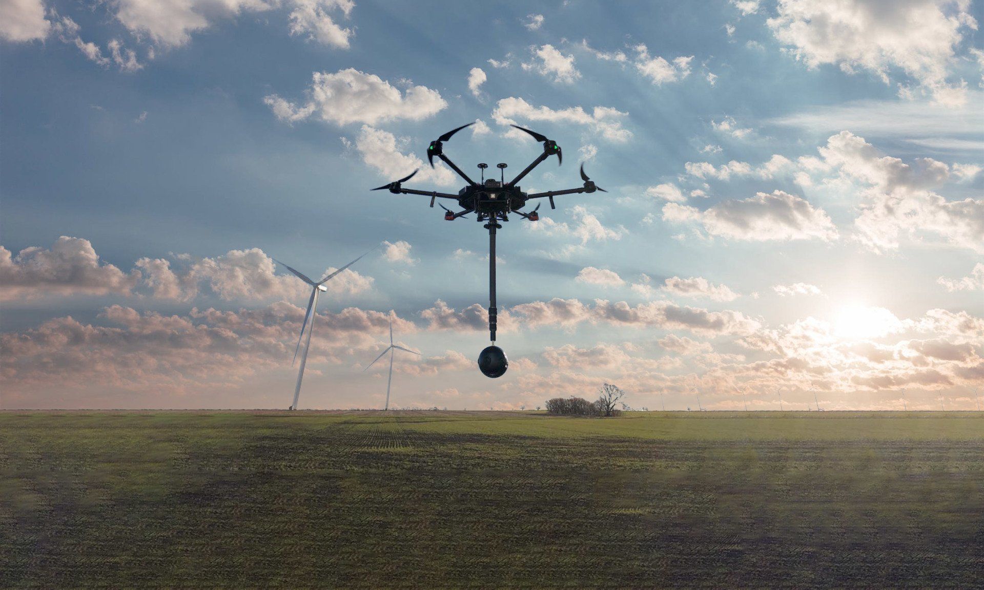 A drone is flying over a field with wind turbines in the background.