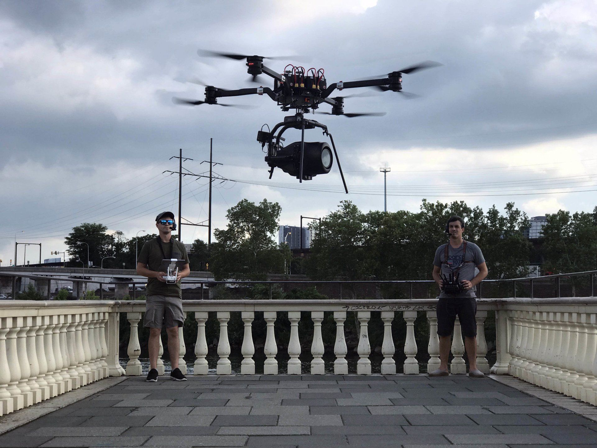 Two men are standing on a balcony watching a drone fly overhead