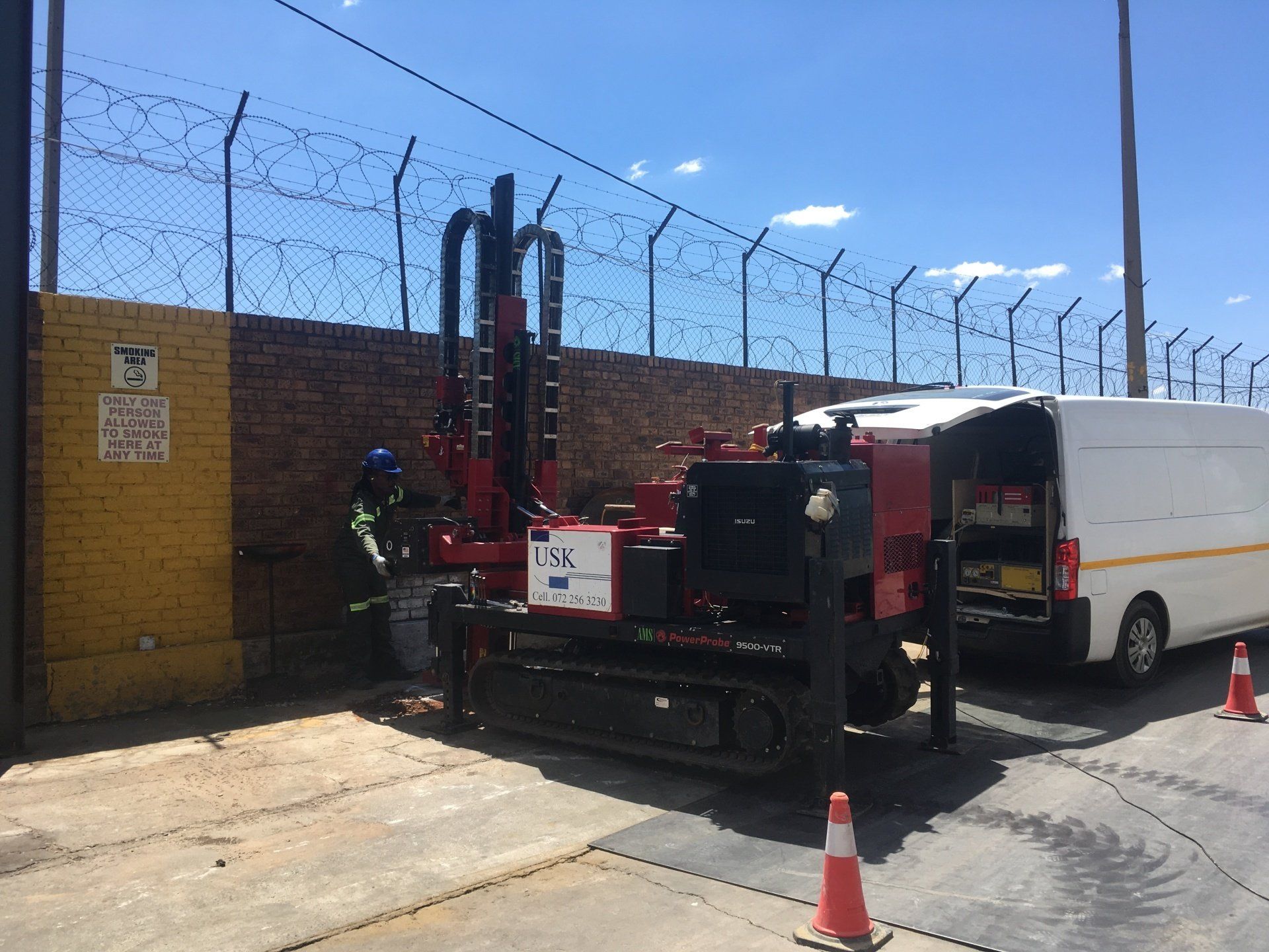 A drilling rig next to a wall, with a worker, white van, and orange cones.