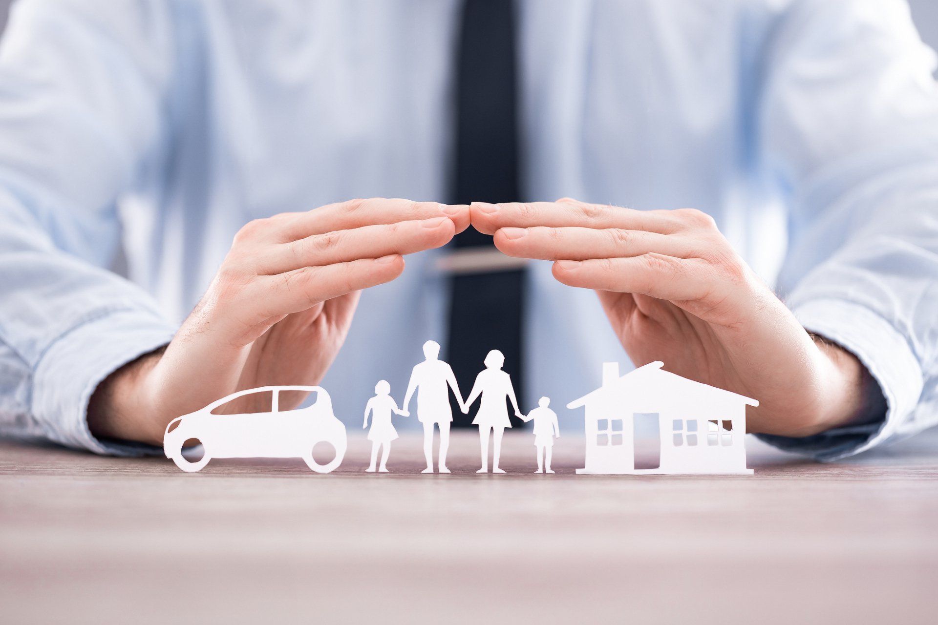 A person in business attire holds their hands protectively over paper cutouts of a car, a family, and a house.