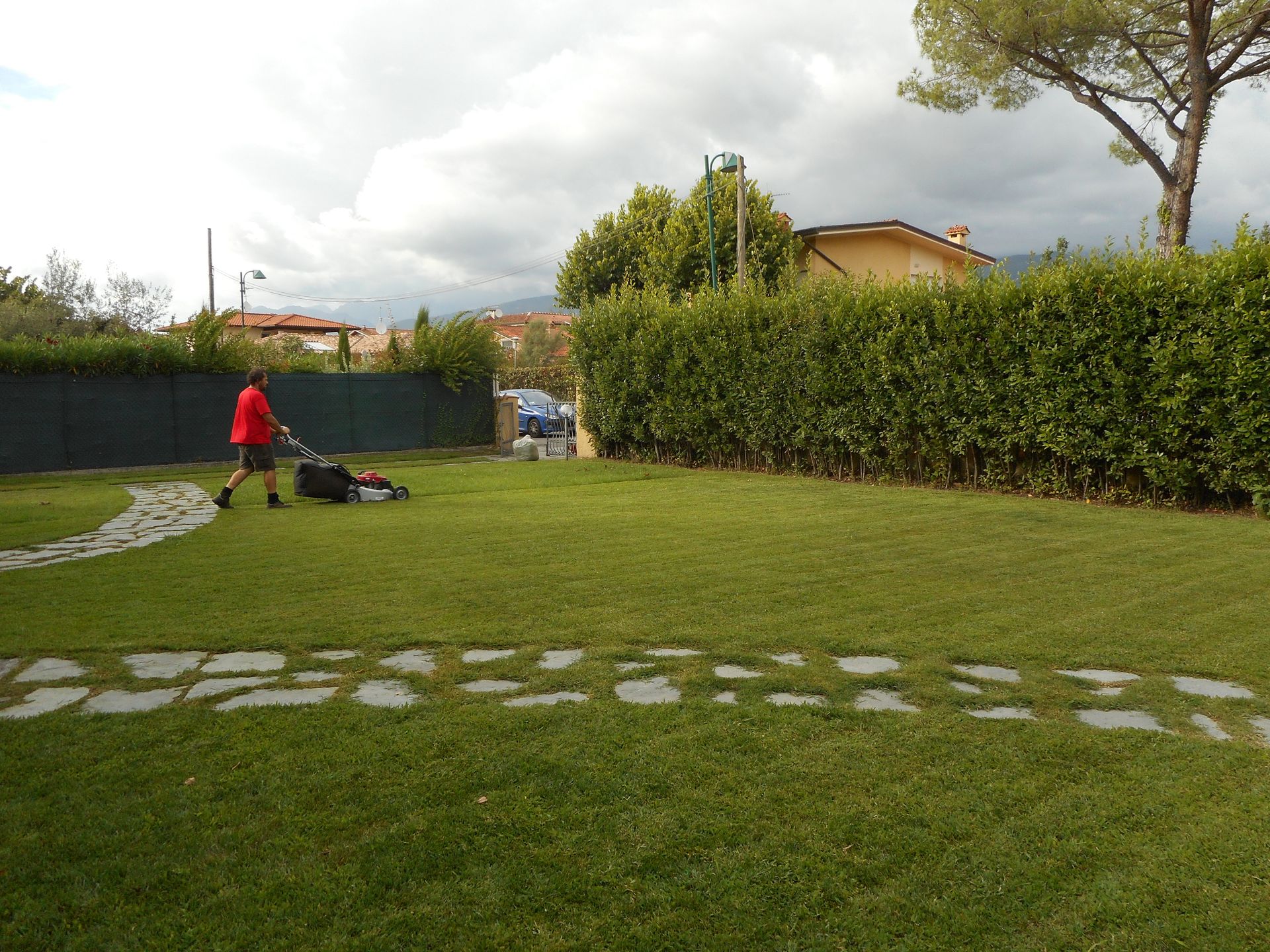 Una persona con una maglietta rossa usa un tosaerba in un giardino verde con un sentiero di pietra sotto un cielo nuvoloso.