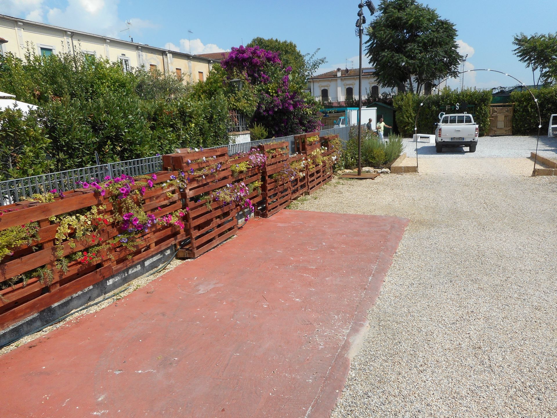 Un sentiero rosso, fiancheggiato da una recinzione di pallet di legno piena di fiori viola, conduce a un parcheggio sterrato dove si trova un camion.