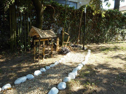 In una giornata di sole, in un cortile si trova una piccola struttura da giardino con il tetto in legno, delimitata da un sentiero fiancheggiato da pietre bianche.