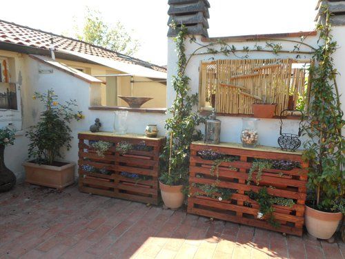 Due fioriere in pallet di legno con piante in vaso sono posizionate su un patio in mattoni, addossate a un muro bianco ricoperto di piante rampicanti.