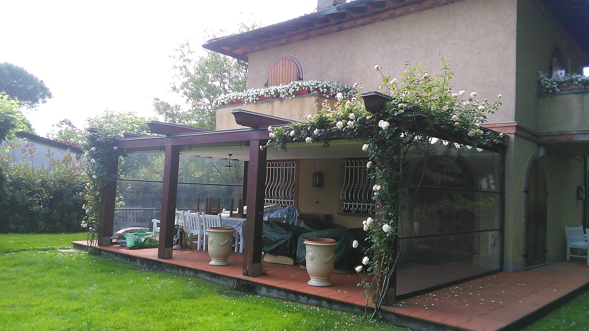 Un pergolato in legno marrone con rose rampicanti bianche copre un patio in terracotta accanto a una casa color sabbia, affacciato su un prato verde.