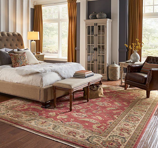 Elegant bedroom with tufted bed, red patterned rug, tall windows, and warm wooden furniture.