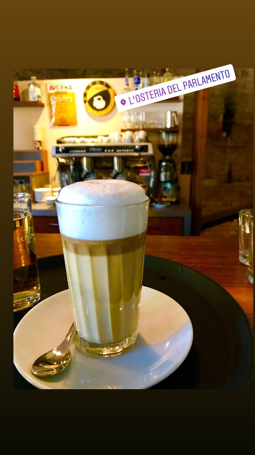 Ein Glas Cappuccino auf einer Untertasse mit einem Löffel auf einem Tisch.
