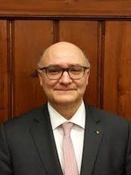 Ein kahlköpfiger Mann mit Brille, Anzug und Krawatte steht vor einer Holzwand.