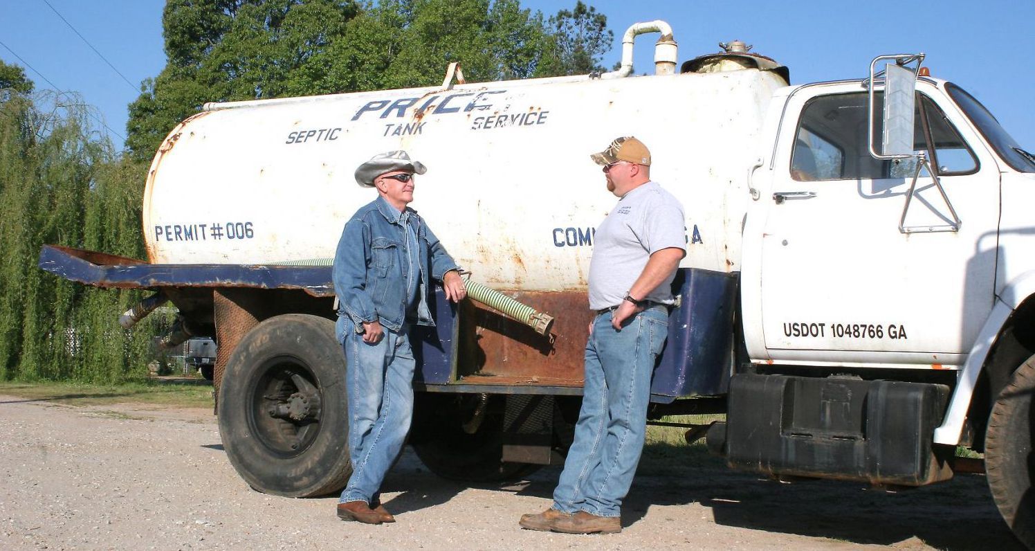 Two men standing next to a water tank truck. One wears a denim jacket and hat. The other wears a ball cap.