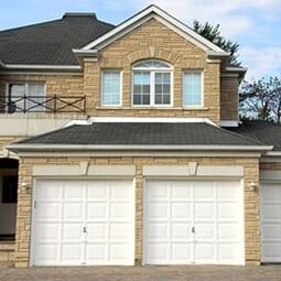 House with garage—General Contractor in Newport News, VA A two-story tan stone house with two white garage doors, a dark shingled roof, and a second-floor arched window.