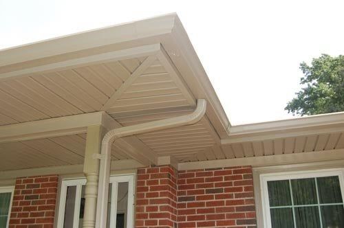 Beige vinyl soffit and fascia under a roof eave, connected to a downspout against a brick house exterior.