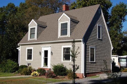 A two-story, shingled house with white trim, two dormer windows, and a brick foundation under a clear blue sky.