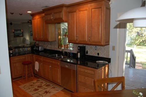 A kitchen with wood cabinets, black countertops, stainless steel appliances, and a floor mat, viewed from a dining area.